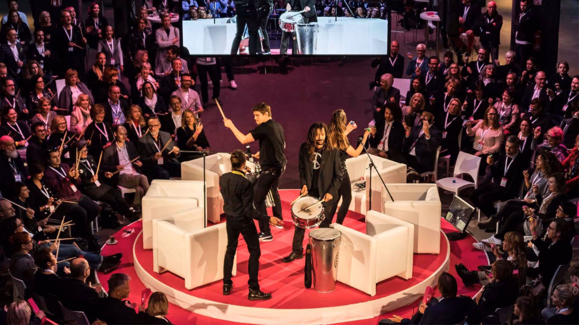 Musiker spielen Schlaginstrumente auf einem runden roten Podium, umgeben von sitzendem Publikum.