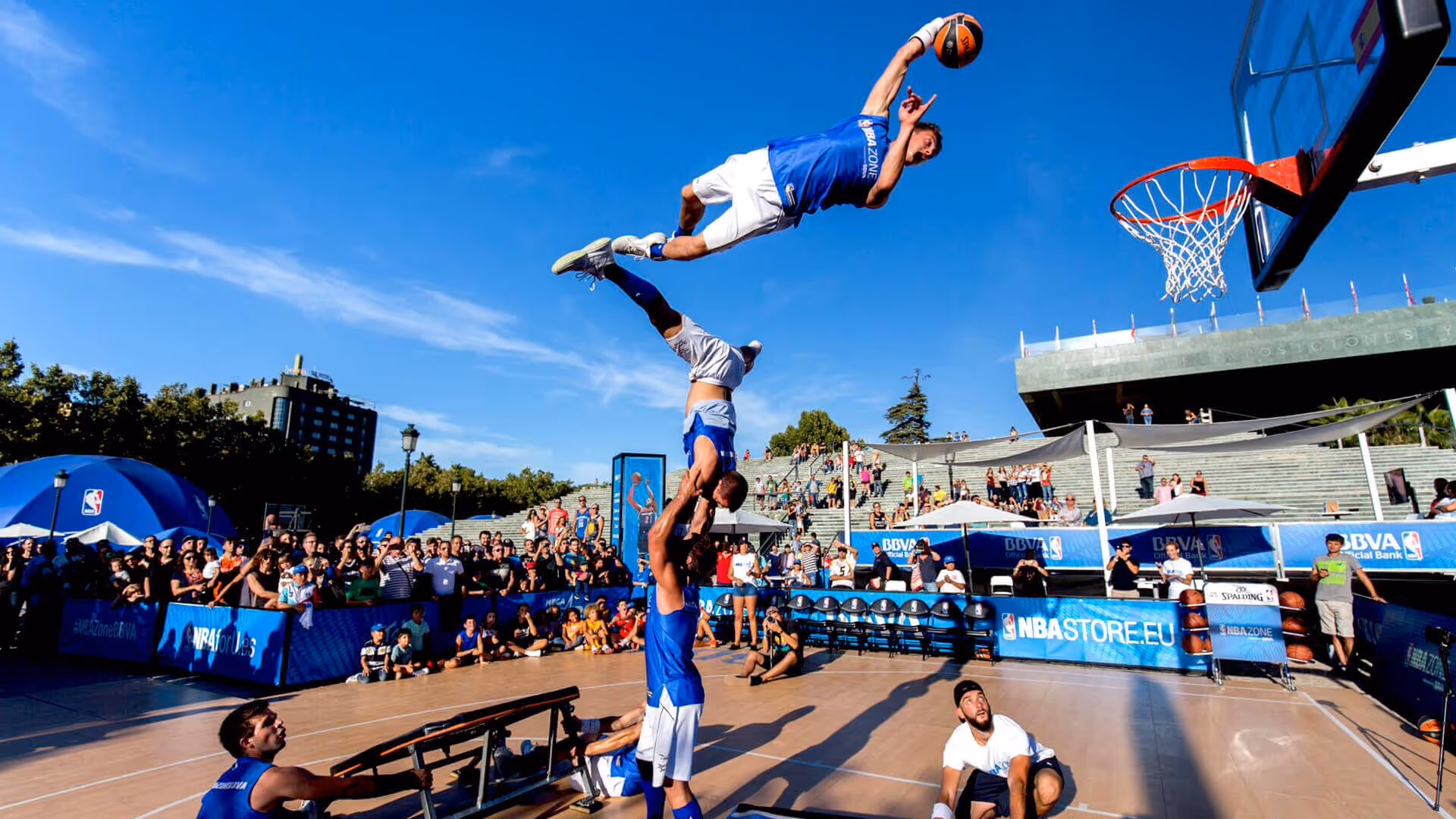 Basketballspieler in blauen Trikots führen einen akrobatischen Dunk durch, wobei ein Spieler einen anderen hochhält, um den Ball in den Korb zu werfen, umringt von einer großen Zuschauergruppe bei einer Outdoor-Veranstaltung.