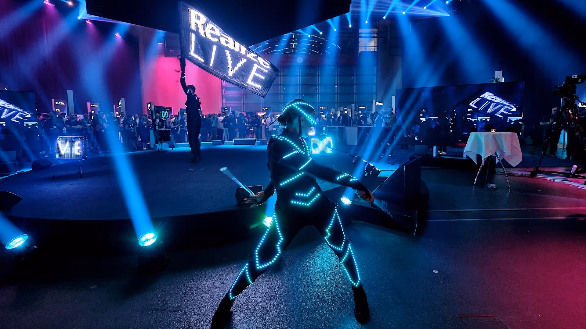 Performance einer Person in einem leuchtenden LED-Anzug auf einer Bühne mit Publikum und blauen und rosa Bühnenlichtern.