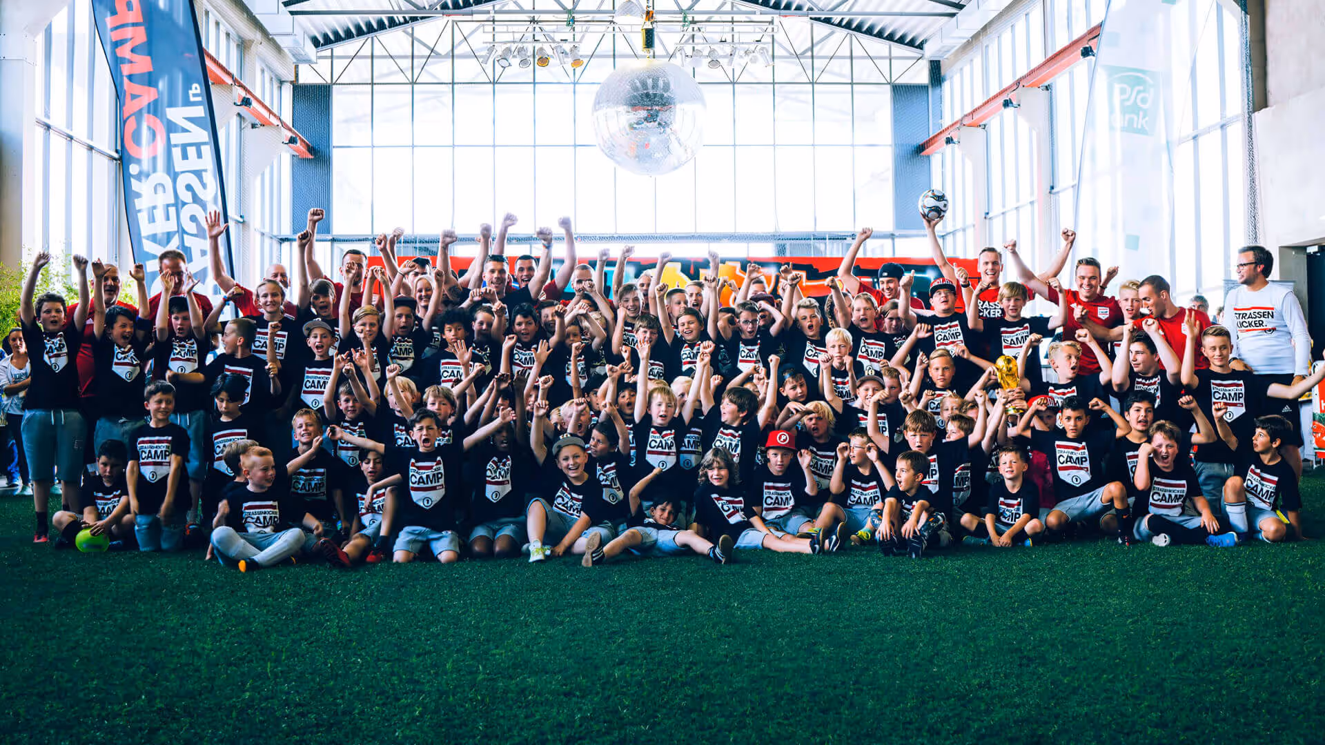 Große Gruppe von Kindern und Erwachsenen in schwarzen T-Shirts mit Aufschrift 'Camp' jubeln bei einem Indoor-Fußballcamp.