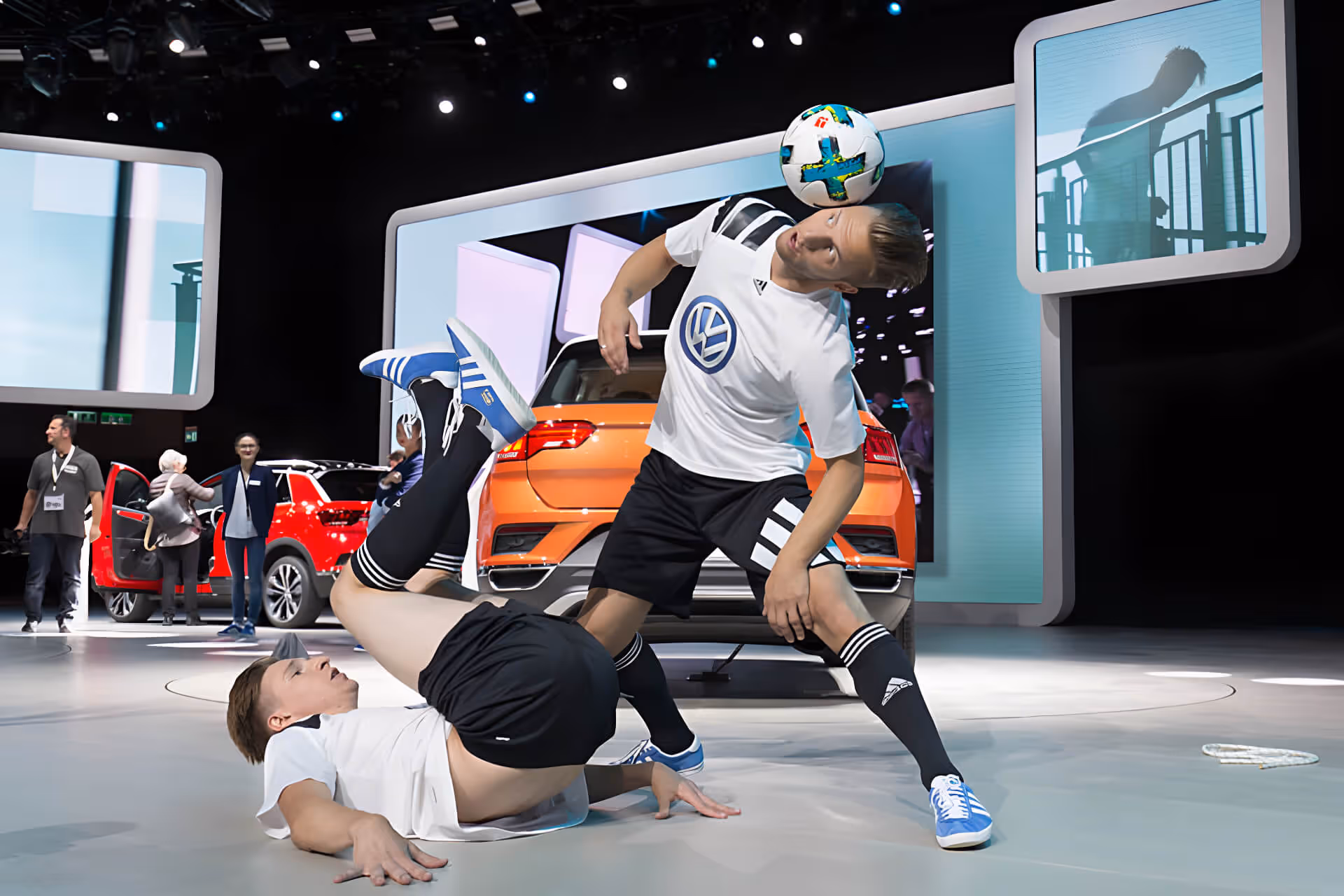 Zwei junge Männer in Fußballausrüstung führen im Showroom vor einem orangen Auto beeindruckende Fußball-Tricks vor.