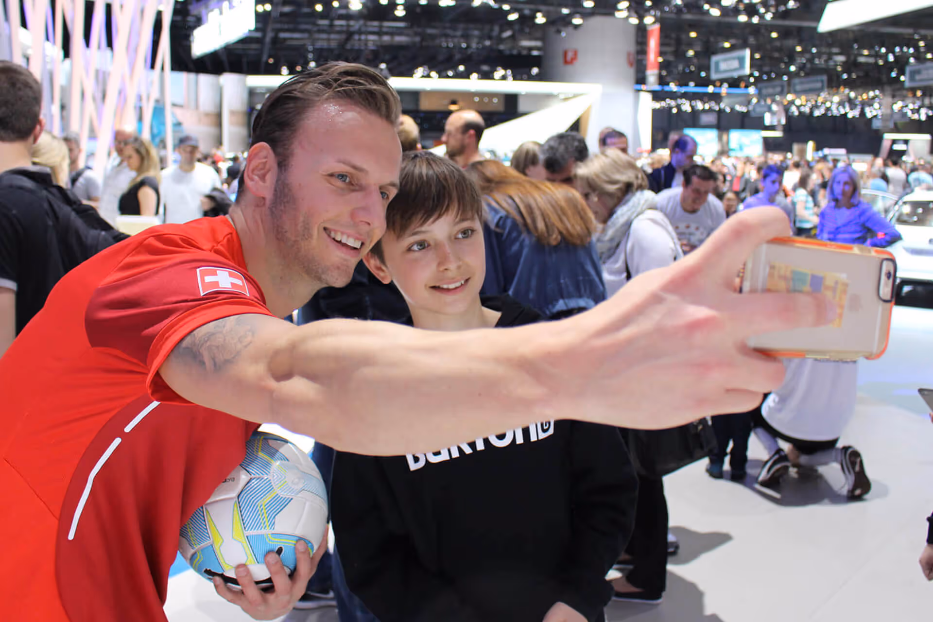 Mann in rotem Trikot mit Schweiz-Flagge macht ein Selfie mit einem Jungen, der einen schwarzen Burton-Hoodie trägt, auf einer belebten Messe.