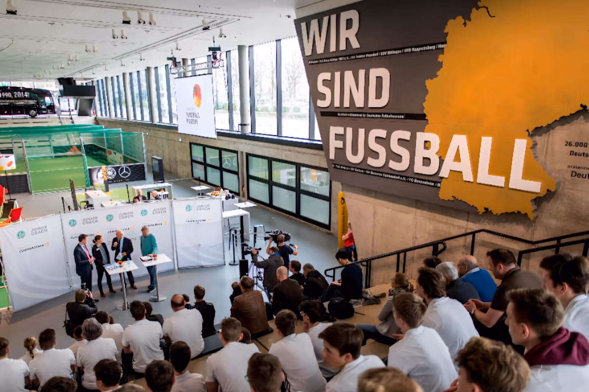 Menschen sitzen auf Tribünen und sehen einer Podiumsdiskussion in einer Sporthalle mit der Schrift ‚Wir sind Fussball‘ an der Wand zu.