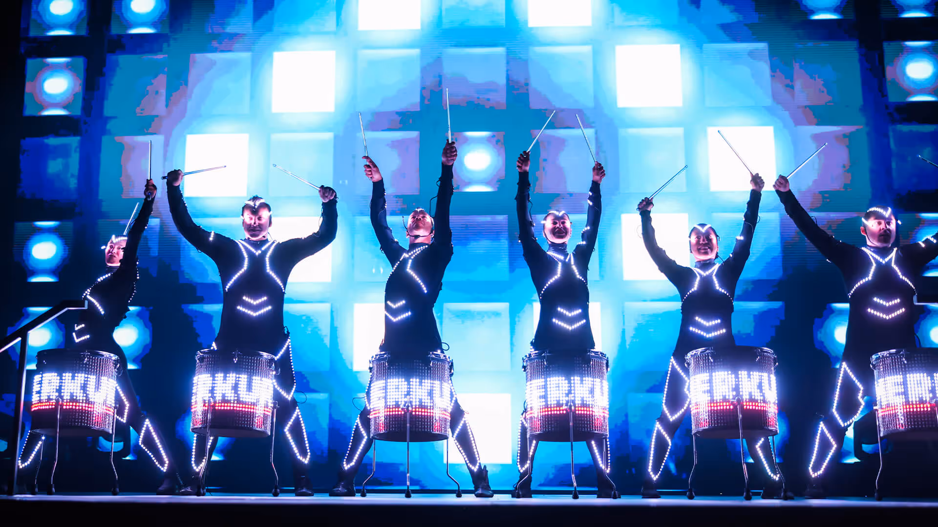 Sechs Trommler in leuchtenden LED-Anzügen spielen vor einer blau-weißen Lichtwand mit erhobenen Trommelstöcken.