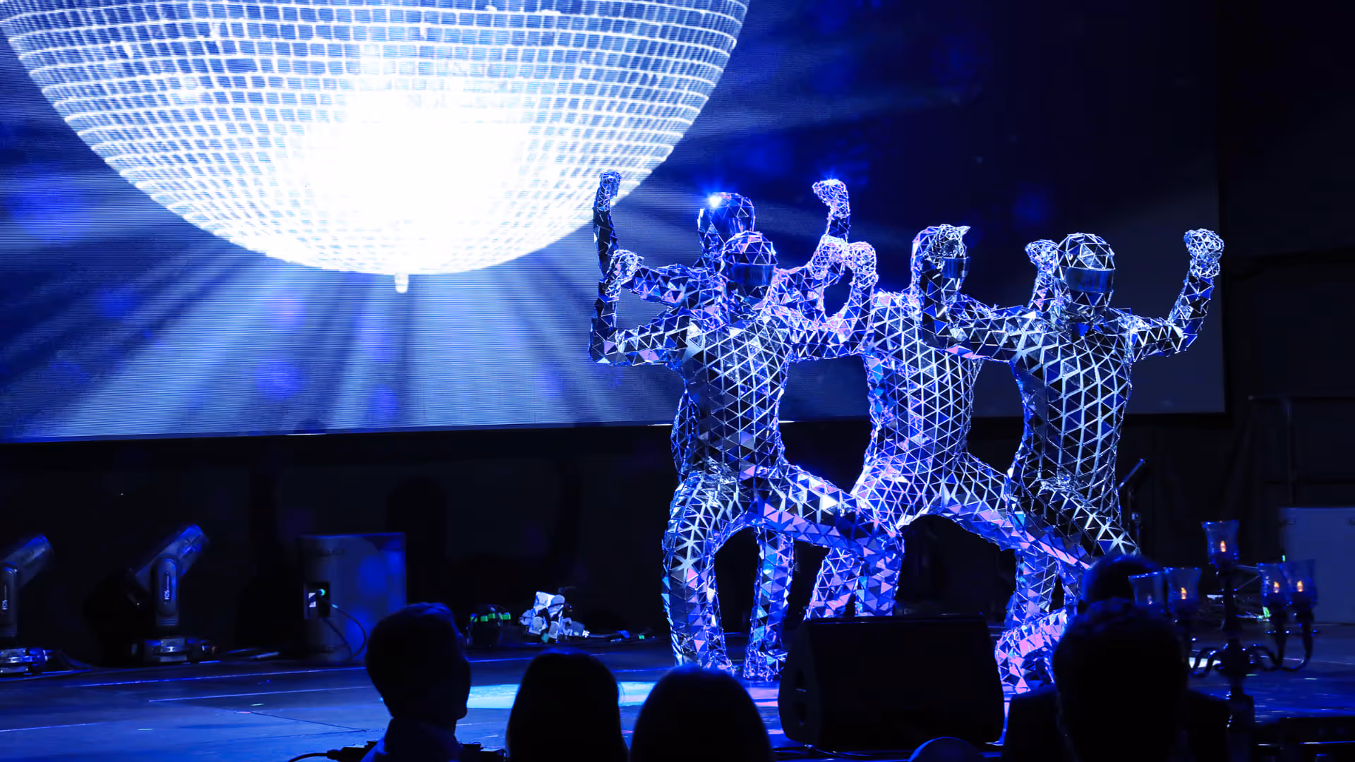 Vier Performer in spiegelnden, geometrischen Anzügen tanzen auf einer Bühne unter einer großen leuchtenden Diskokugel.