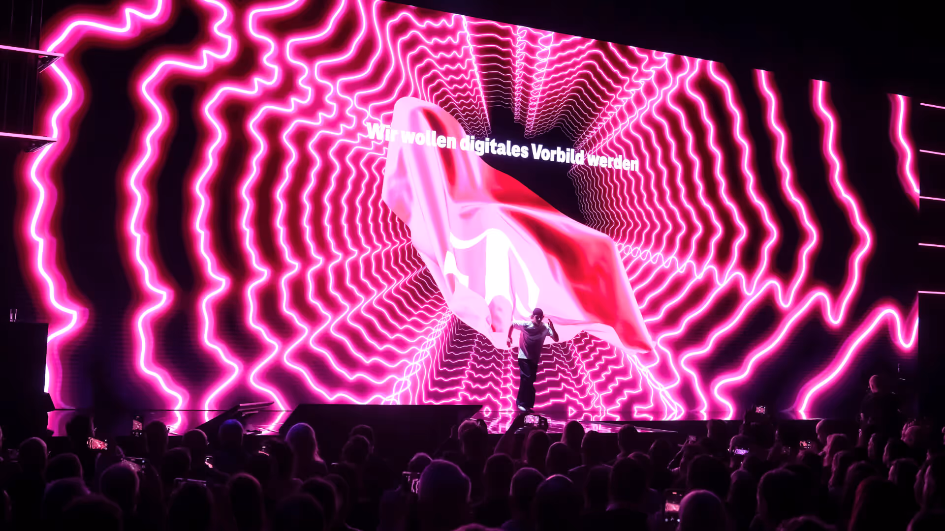 Person auf Bühne vor großer LED-Leinwand mit pinken, wellenförmigen Lichtern und Text "Wir wollen digitales Vorbild werden".