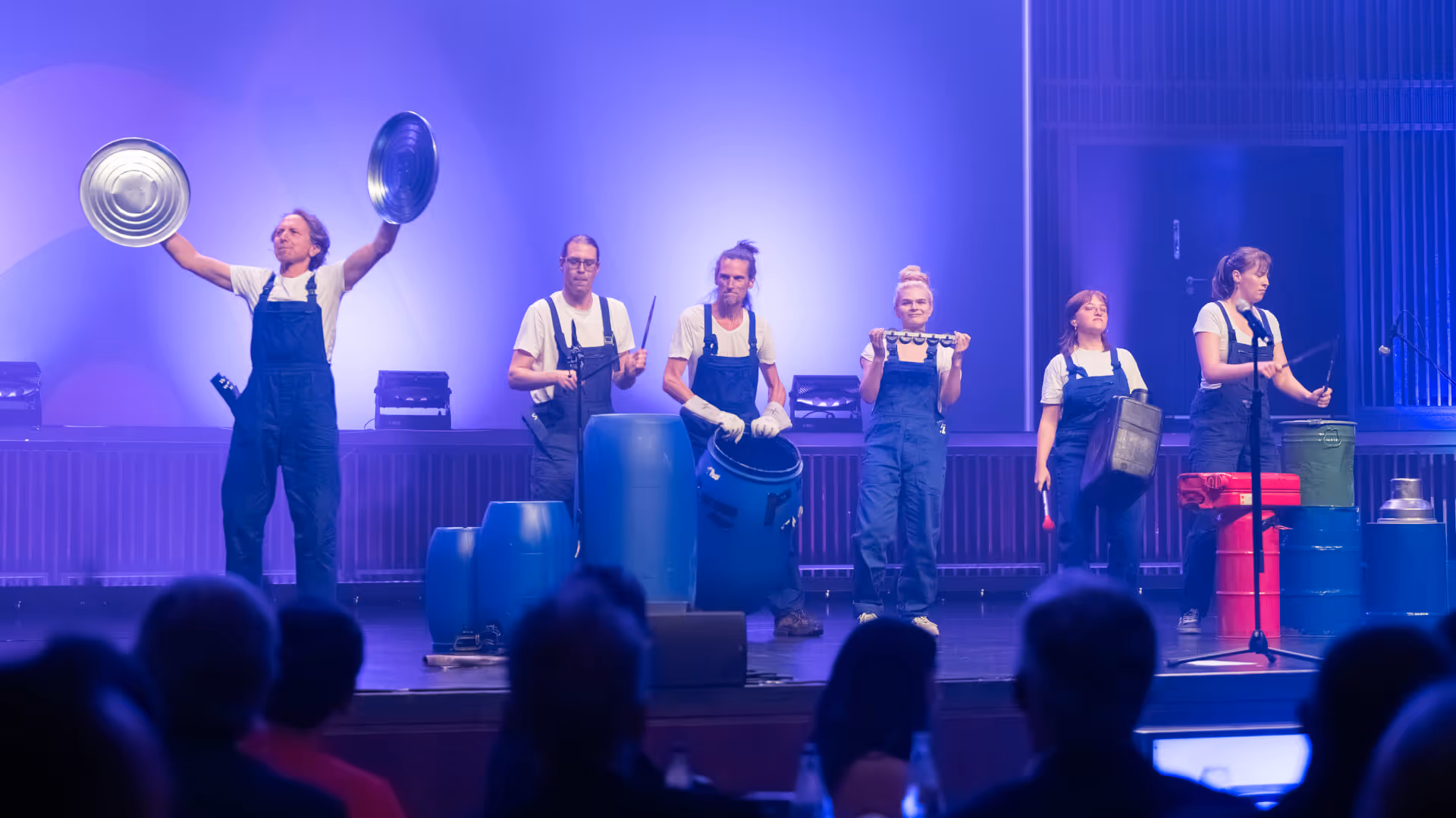 Sechs Personen in blauen Overalls spielen auf der Bühne auf improvisierten Schlaginstrumenten wie Eimern und Metallplatten vor einem Publikum.