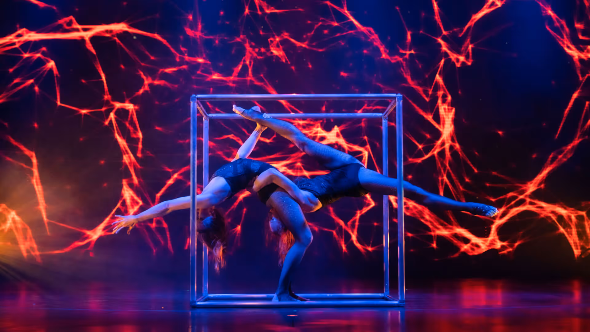 Zwei Akrobatinnen in schwarzen Outfits führen eine komplexe Pose zusammen in einem metallischen Rahmen vor einem Bühnenhintergrund mit roten, leuchtenden Linien aus.