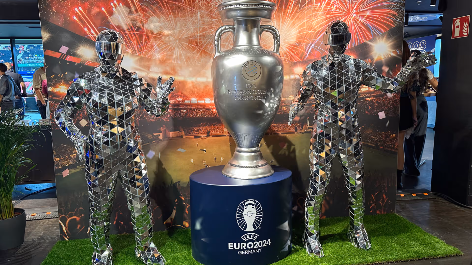 Zwei Spiegelkostüm-Figuren posieren neben dem UEFA EURO 2024 Pokal auf einem Podest mit grünem Kunstrasen und Feuerwerk im Stadion-Hintergrund.