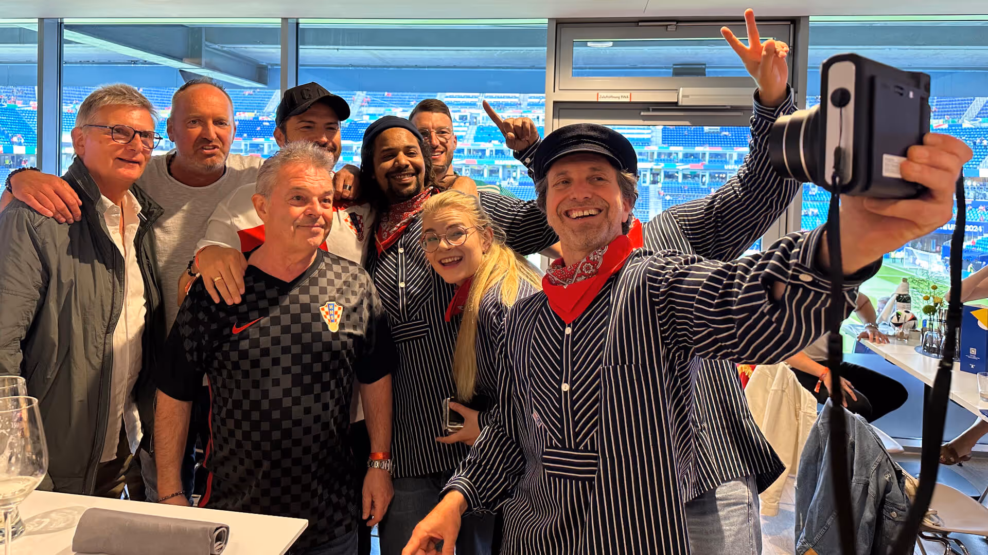 Gruppe fröhlicher Menschen in einem Stadion nimmt ein Selfie, einige tragen schwarz-weiß gestreifte Kleidung und rote Halstücher.