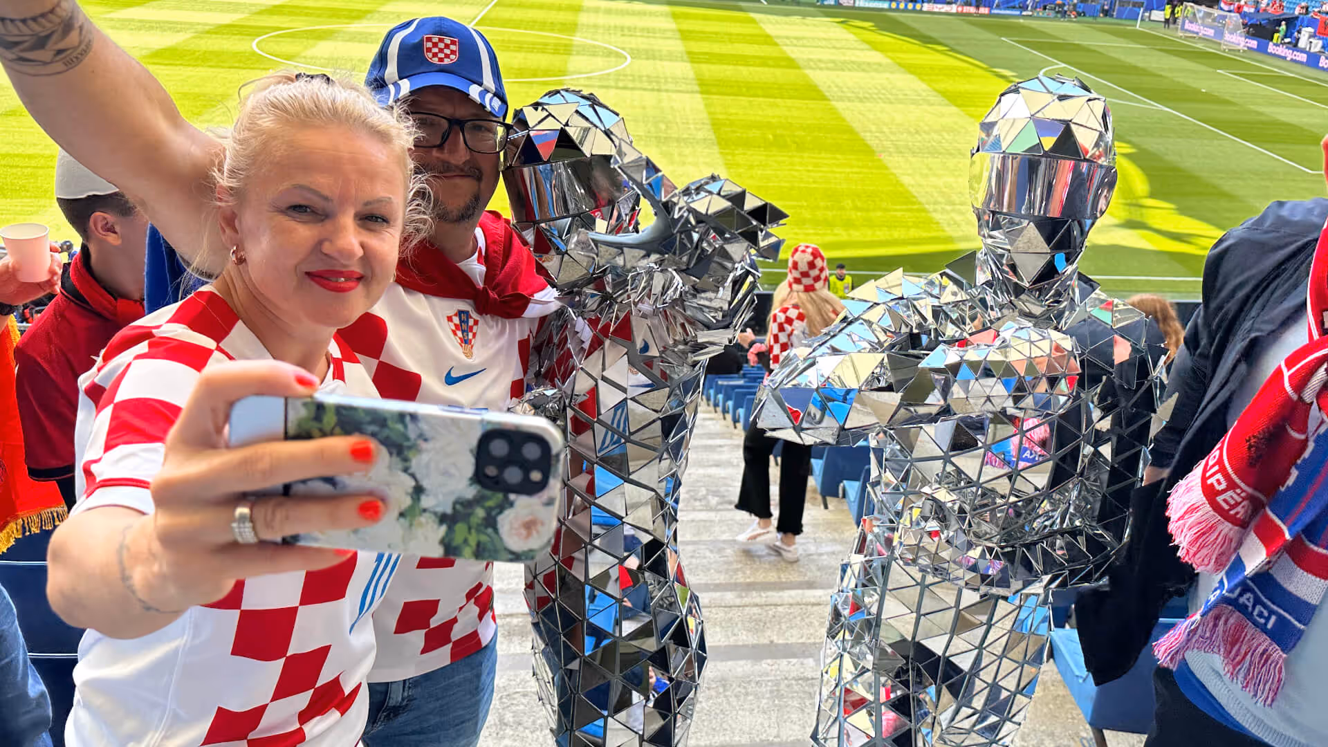 Zwei Fans in kroatischen Fußballtrikots machen ein Selfie mit zwei Personen in glänzenden Spiegelanzügen im Stadion.