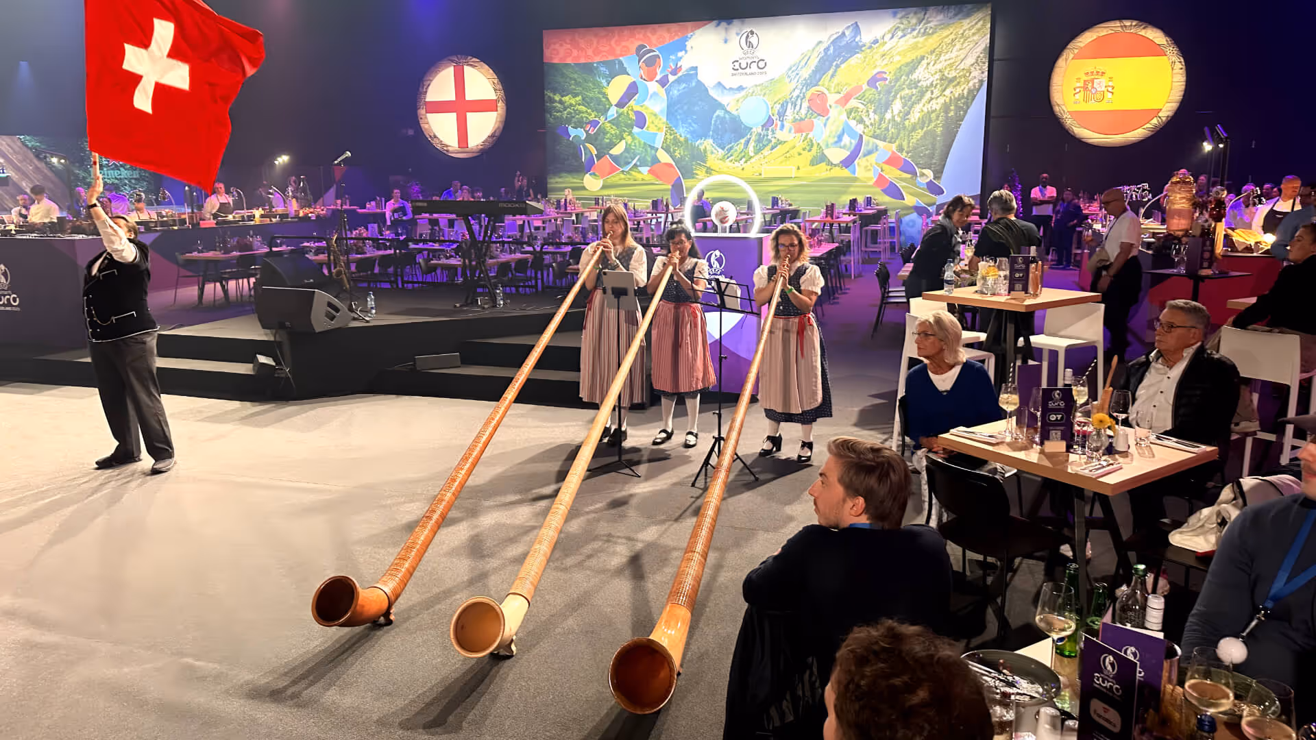 Drei Frauen in traditioneller Tracht spielen Alphörner in einer Veranstaltungshalle, während ein Mann mit Schweizer Fahne daneben steht.