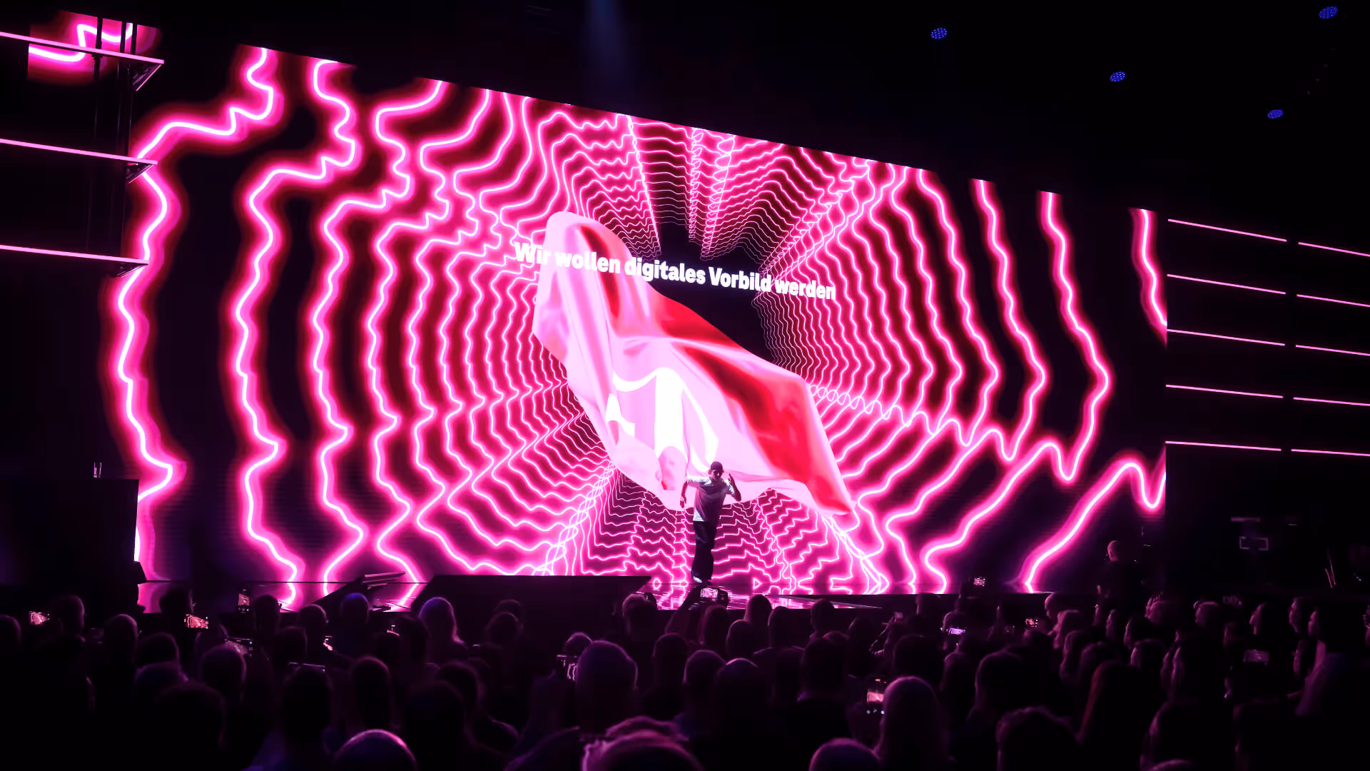 Person auf Bühne vor großem Bildschirm mit pinken, wellenförmigen Neonlinien, der Text 'Wir wollen digitales Vorbild werden' und einer wehenden roten Fahne.