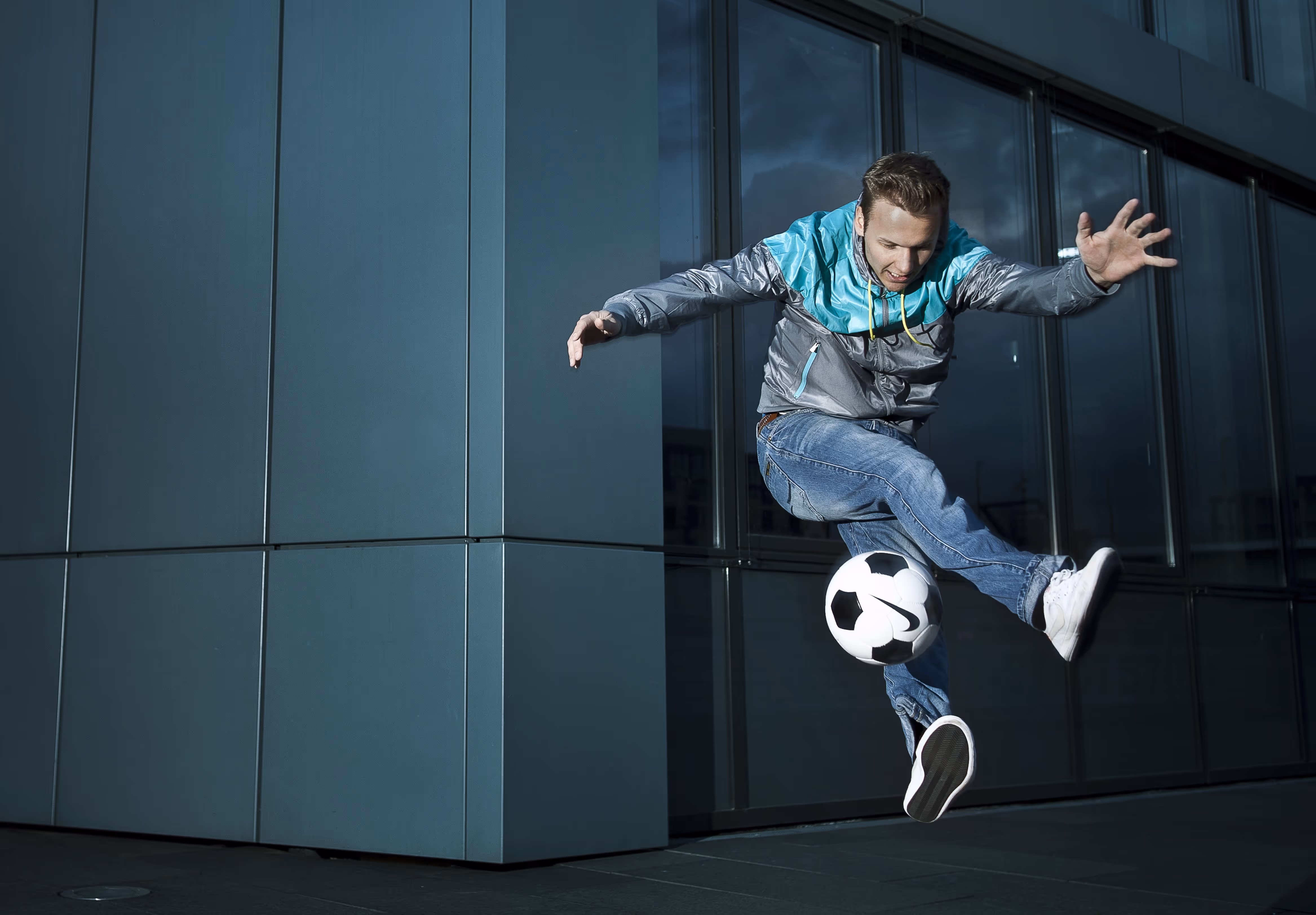Ein Mann in Jeans und winddichter Jacke spielt Fußball vor einem modernen Glasgebäude.