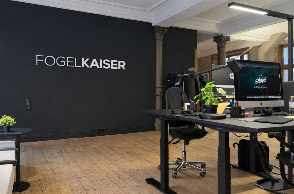 Modernes Büro mit schwarzer Wand und dem Schriftzug FOGELKAISER, Arbeitsplätze mit Computern und Pflanzen auf Holzboden.