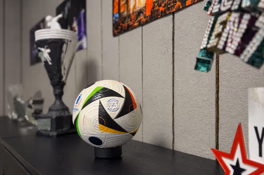 Ein Fußball steht auf einem schwarzen Sockel auf einem schwarzen Möbelstück, daneben ein großer Pokal und weitere Auszeichnungen an der Wand.