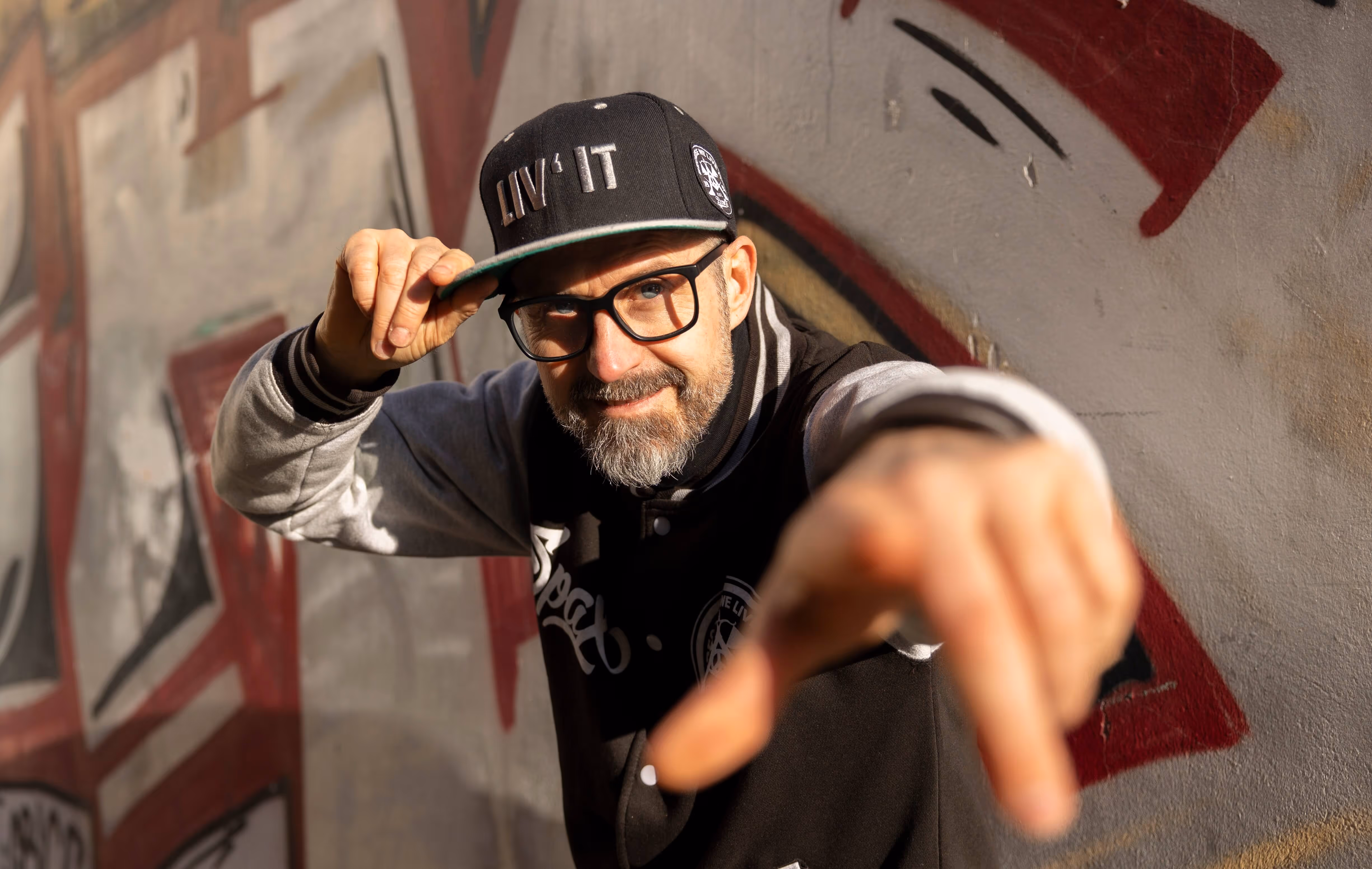 Rapper mit Brille vor einer Wand zeigt in die Kamera