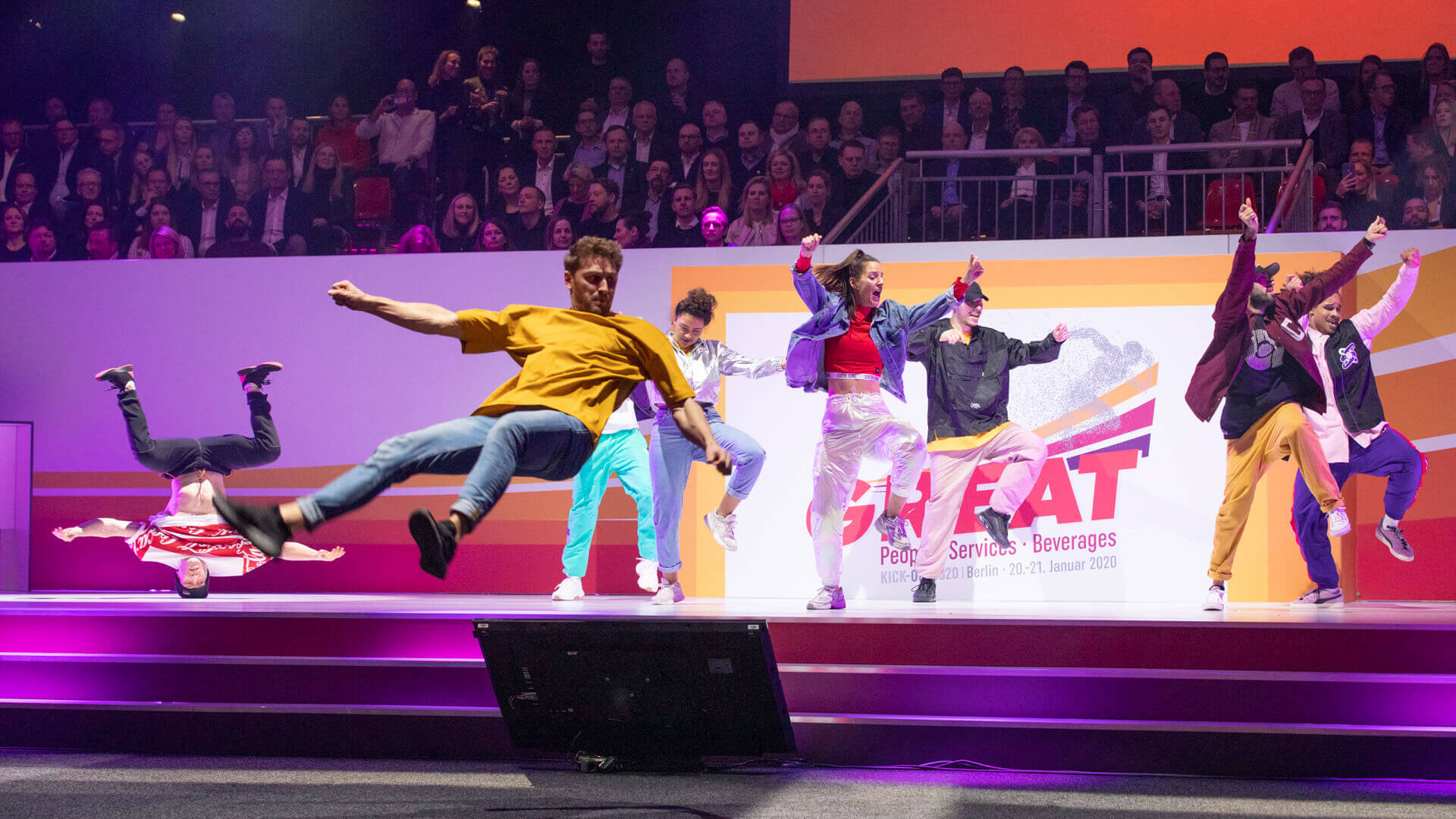 Tanzgruppe bei einer Bühnenshow: mehrere Tänzer springen und machen akrobatische Moves vor Publikum in einer Eventhalle.