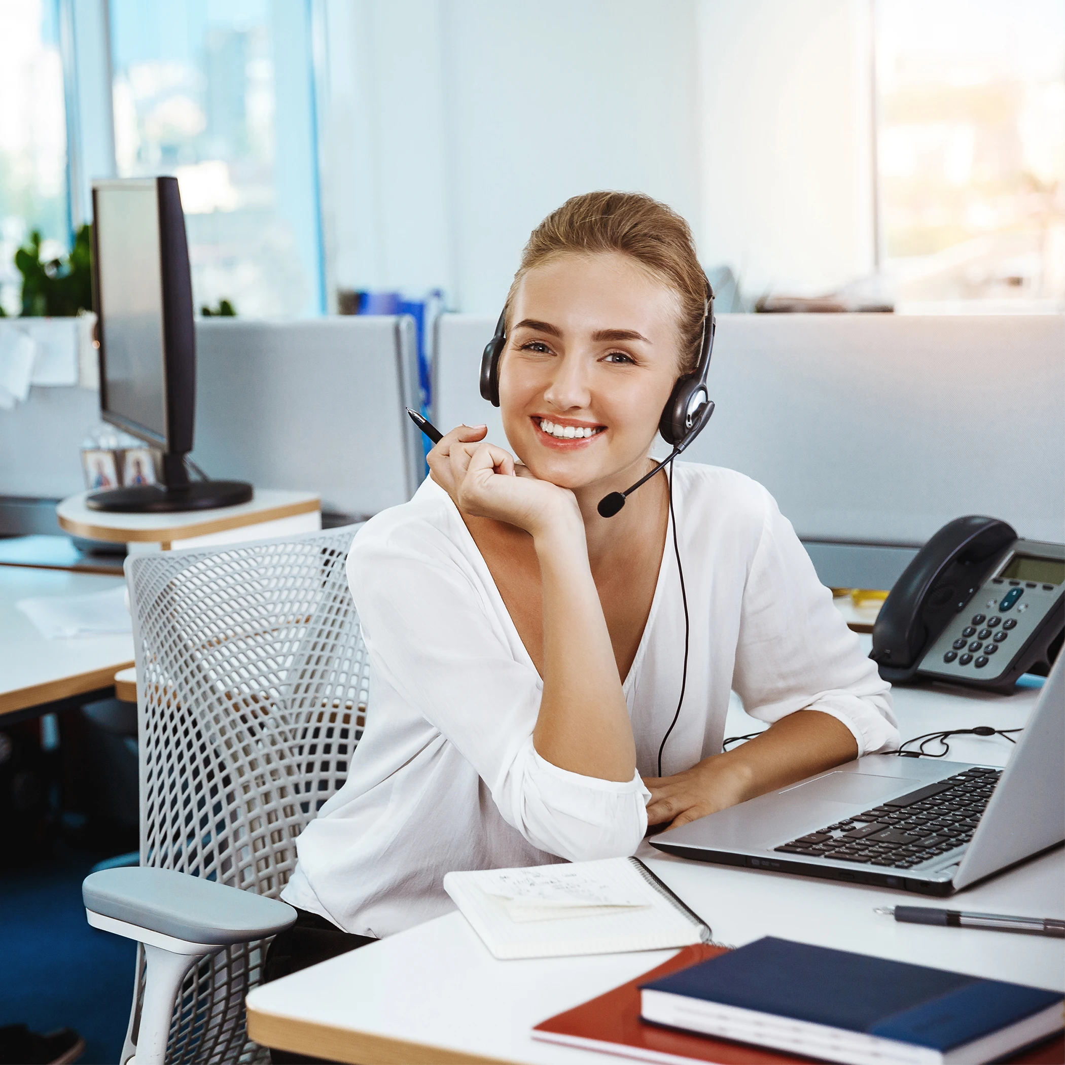Vrouwelijke medewerker van ClikQ met een headset glimlacht en zit bij een bureau met laptop en notitieboekjes in een kantooromgeving.