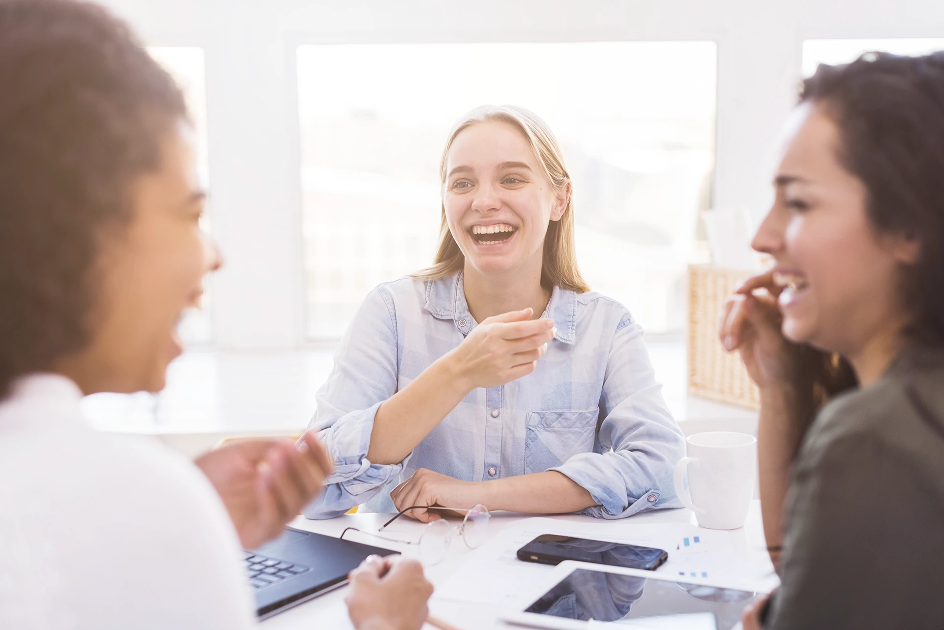 Drie vrouwelijke medewerkers van ClikQ lachen en hebben een vrolijk gesprek aan een tafel met laptop, telefoon en documenten.