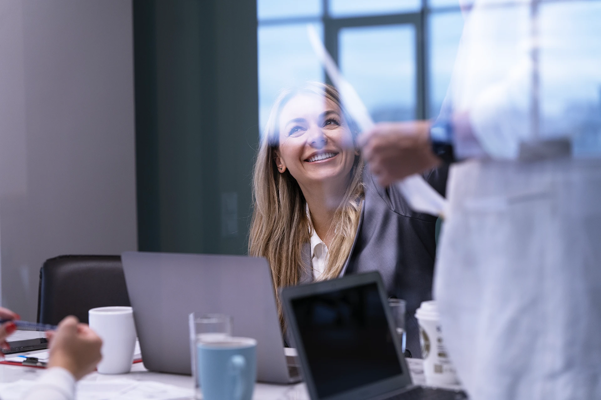 Vrouwelijke collega van ClikQ die glimlacht naar een staande collega tijdens een vergadering aan een tafel met laptops en koffiekopjes.