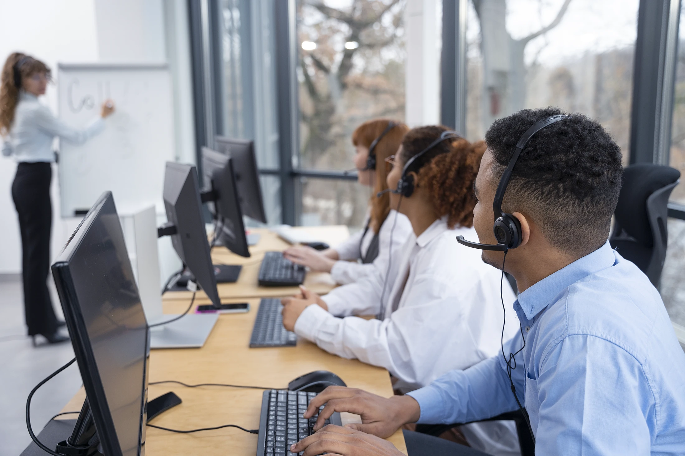 Drie collega’s van ClikQ met headset zitten achter computers en volgen een training van een vrouw die op een whiteboard schrijft.