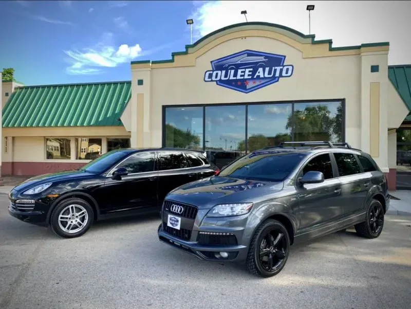 Two luxury SUVs, a black Porsche and a grey Audi, parked in front of the Coulee Auto dealership on a clear day