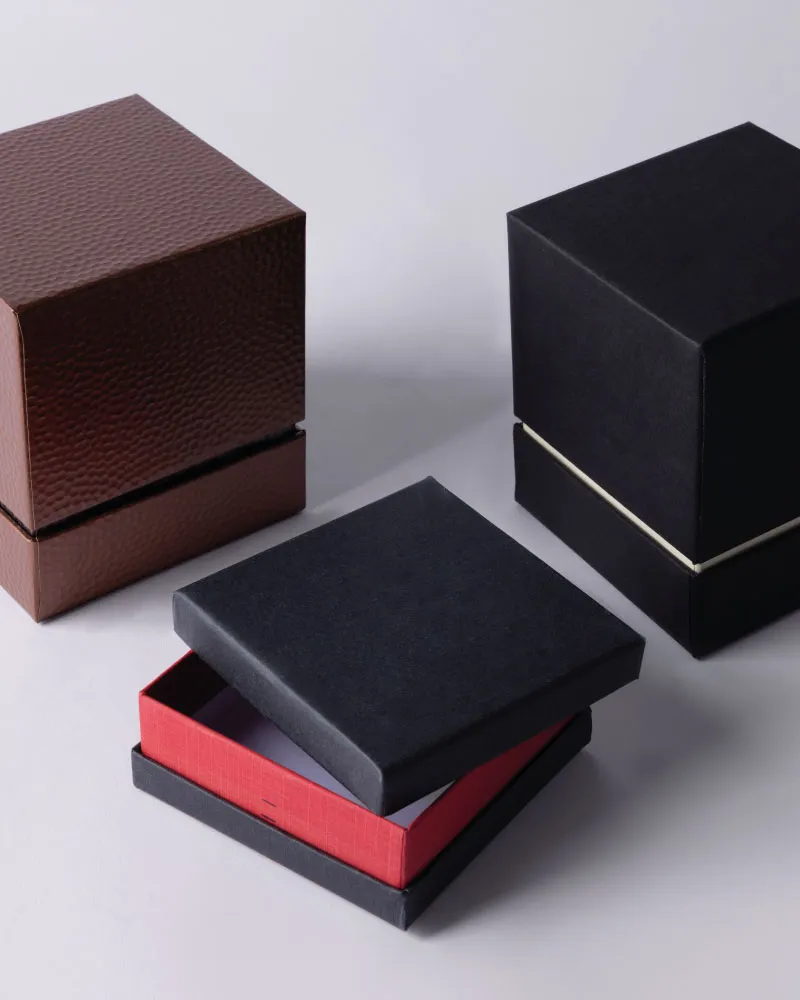 Three rigid packaging boxes including a brown textured closed box, a black closed box with a thin gold band, and a black box with a red interior partially open.