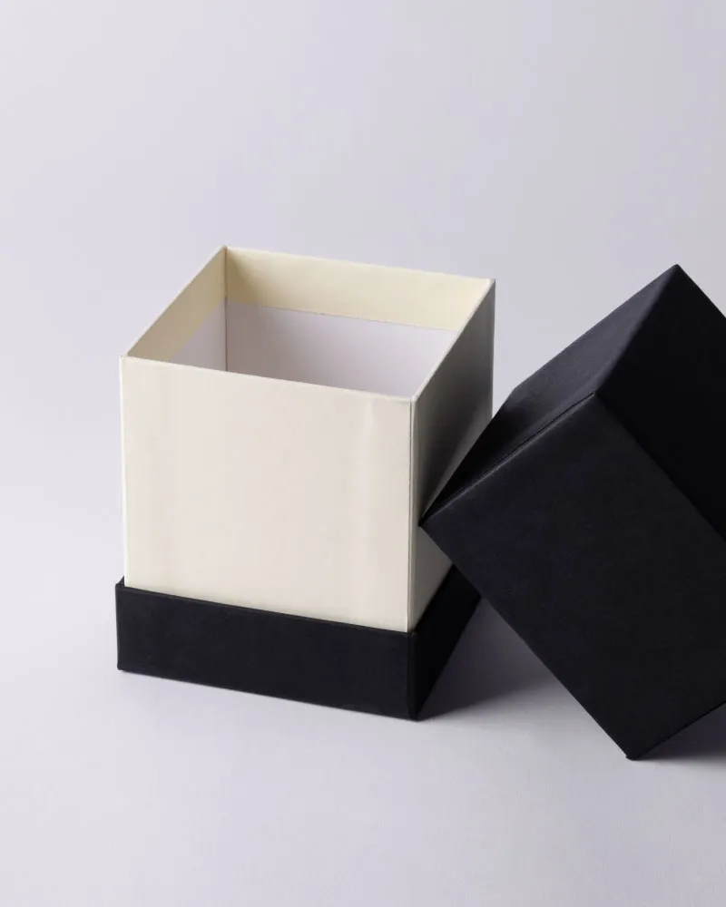 Open square rigid box with a cream-colored inner section and black base, next to its detached black lid.