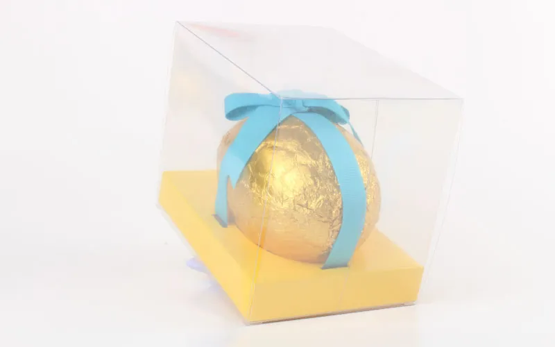 Gold foil-wrapped egg with a blue ribbon inside a clear plastic box with a yellow base.