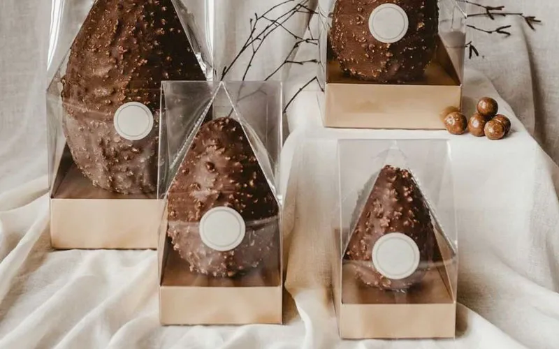 Four chocolate eggs with textured surfaces, each in clear plastic packaging with gold bases, arranged on a white fabric.
