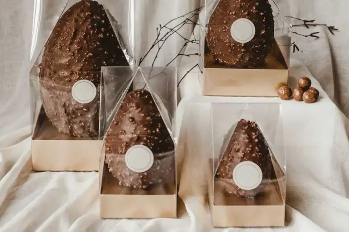 Four chocolate Easter eggs with nuts, each in a clear plastic triangular box with a golden base and a round white label.