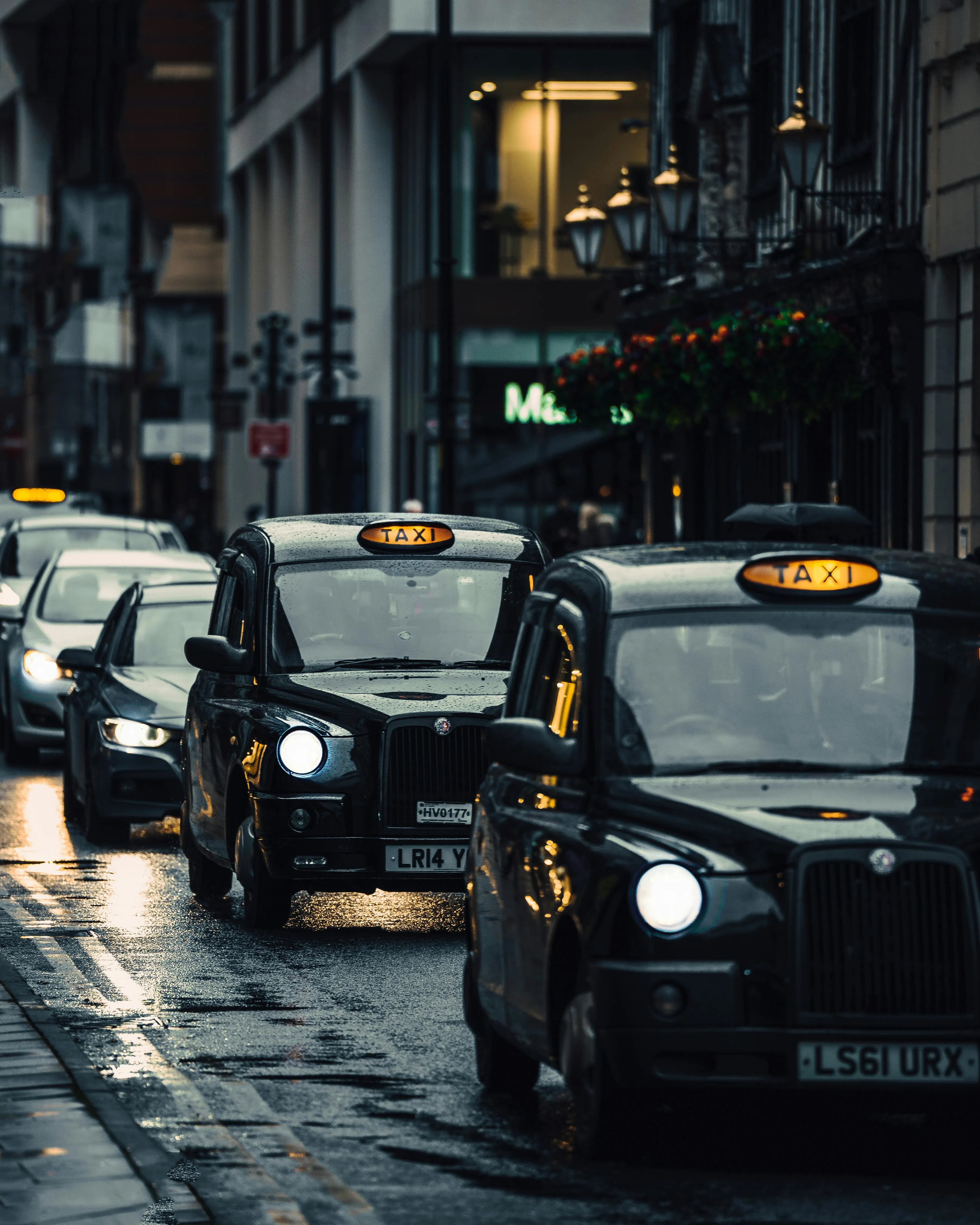 Black London taxis lined up on a wet, dimly lit city street at dusk