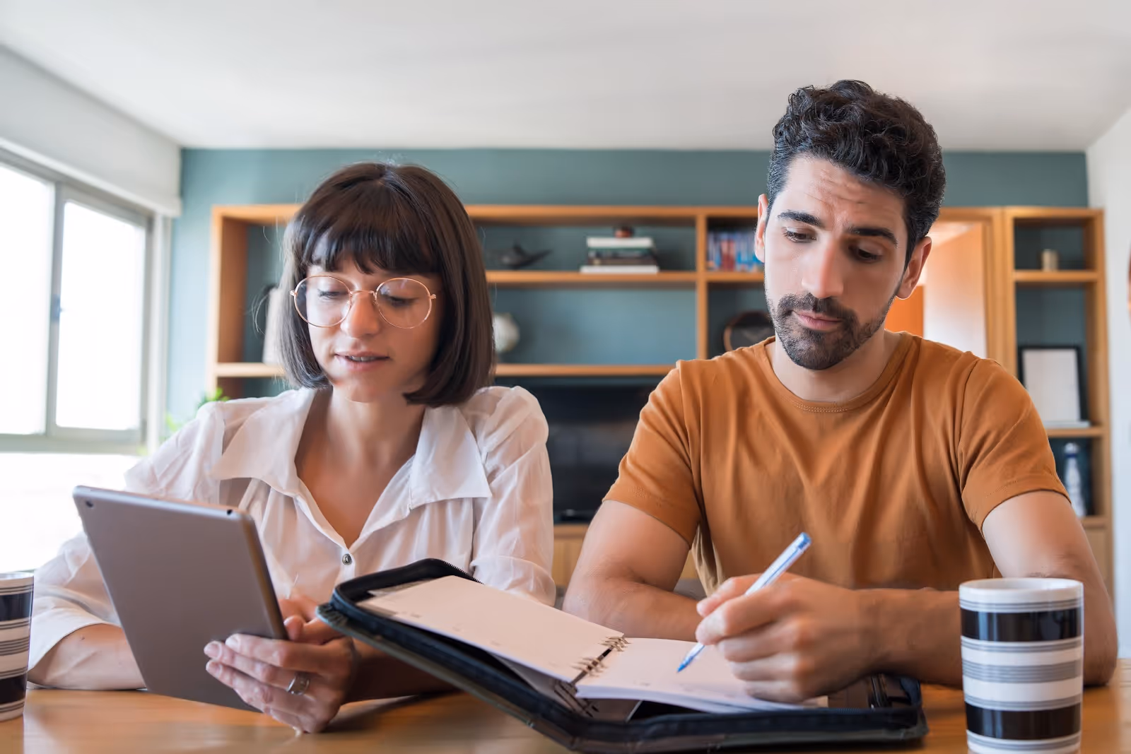 Man and woman sitting at a table reviewing notes, with the woman holding a tablet and the man writing in a notebook.
