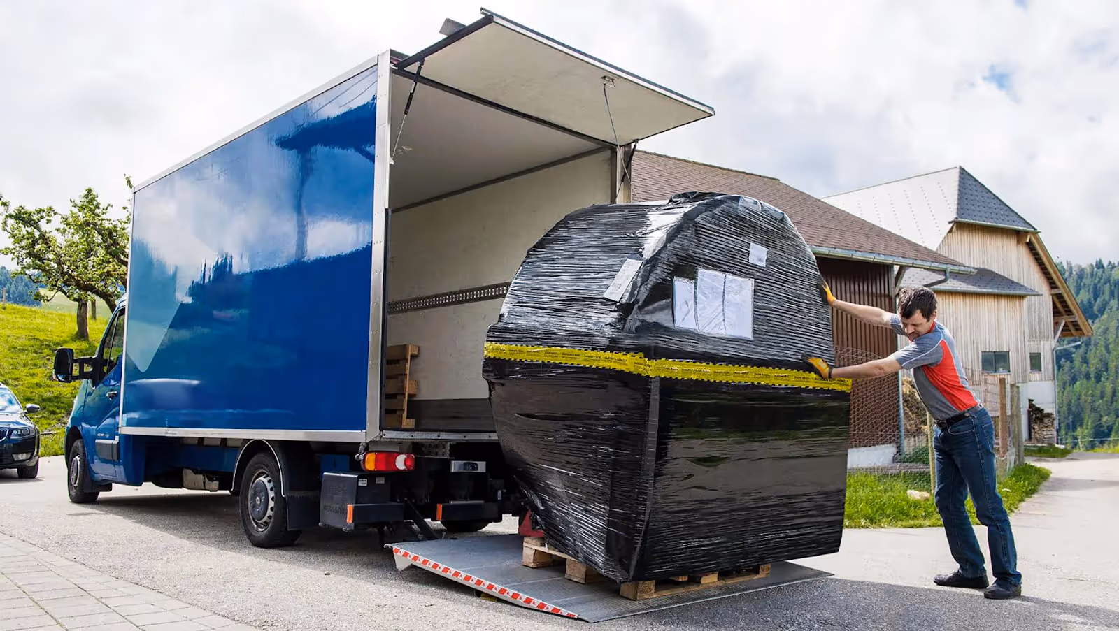 Man pushing a large black wrapped pallet onto the back of a blue delivery truck using a metal ramp.