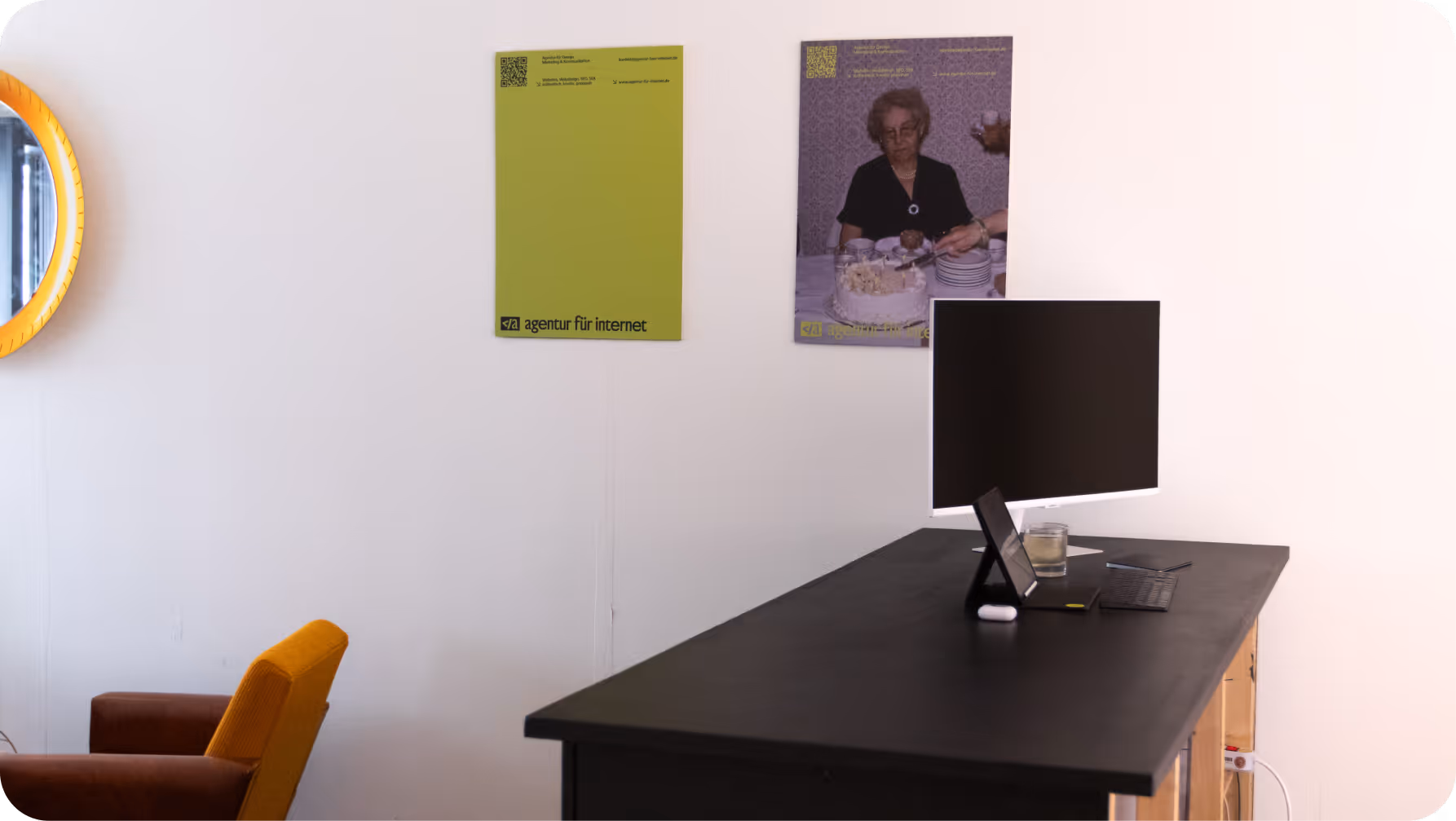Moderner Arbeitsbereich mit schwarzem Tisch, Monitor, gelbem Stuhl, Spiegel und zwei Wandpostern.