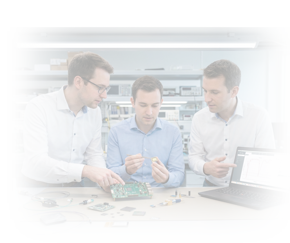 Three engineers in a lab examining a circuit board and a component while discussing data shown on a laptop screen.