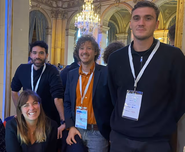 Quatre personnes souriantes avec des badges, dans un décor avec un grand lustre et des colonnes ornées.
