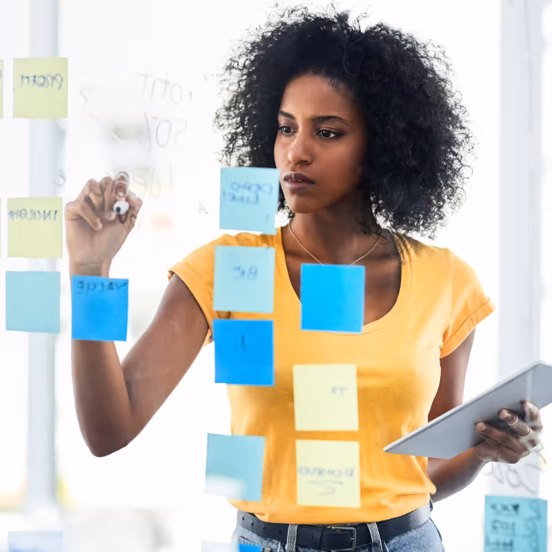 Femme concentrée écrivant des notes sur un tableau en verre avec des post-its colorés, tenant une tablette.