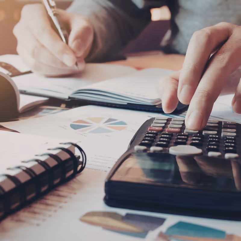 Une main utilise une calculatrice tandis qu’une autre écrit dans un carnet sur une table avec des documents.