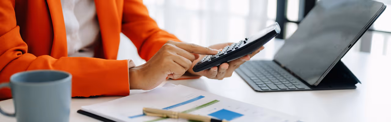 Person in orange jacket using calculator near tablet and financial documents on white desk.
