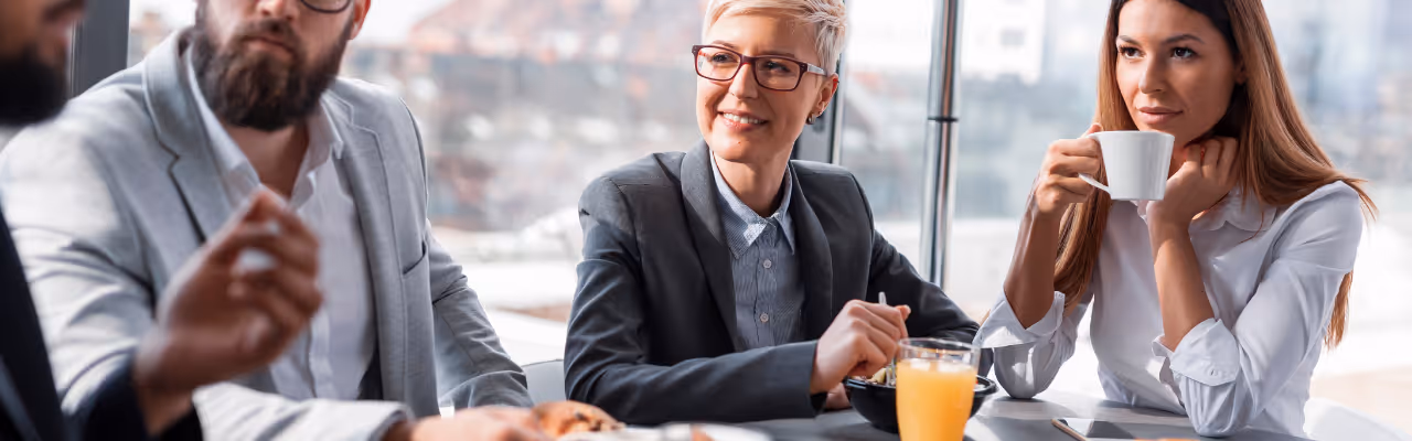 Trois collègues discutent autour d'une table lors d'une pause déjeuner en salle de réunion.