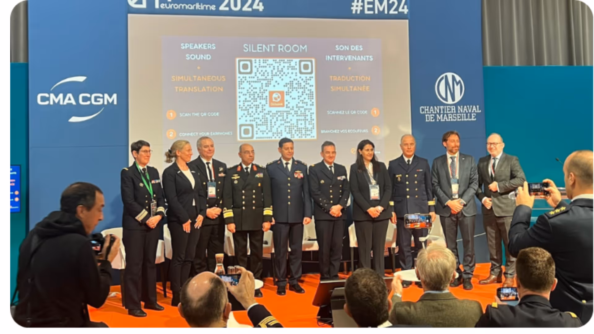 Groupe de professionnels en uniforme posant devant un écran montrant un QR code lors d'un événement maritime.