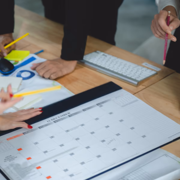 Des mains de plusieurs personnes autour d'un calendrier mensuel sur un bureau en bois, avec un clavier, stylos et autres fournitures de bureau visibles.