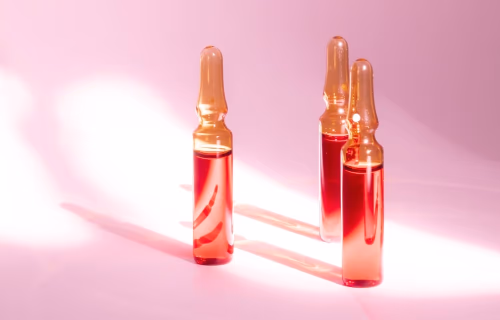 Three amber glass ampoules filled with red liquid on a pink surface with soft lighting.