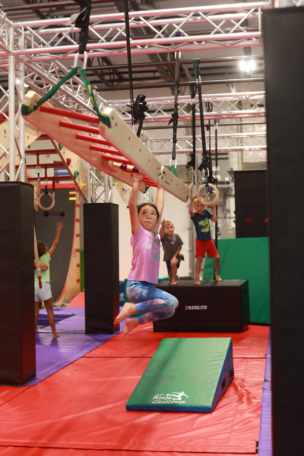 girl confidently swinging across hanging ladder while other ninjas waiting their turn