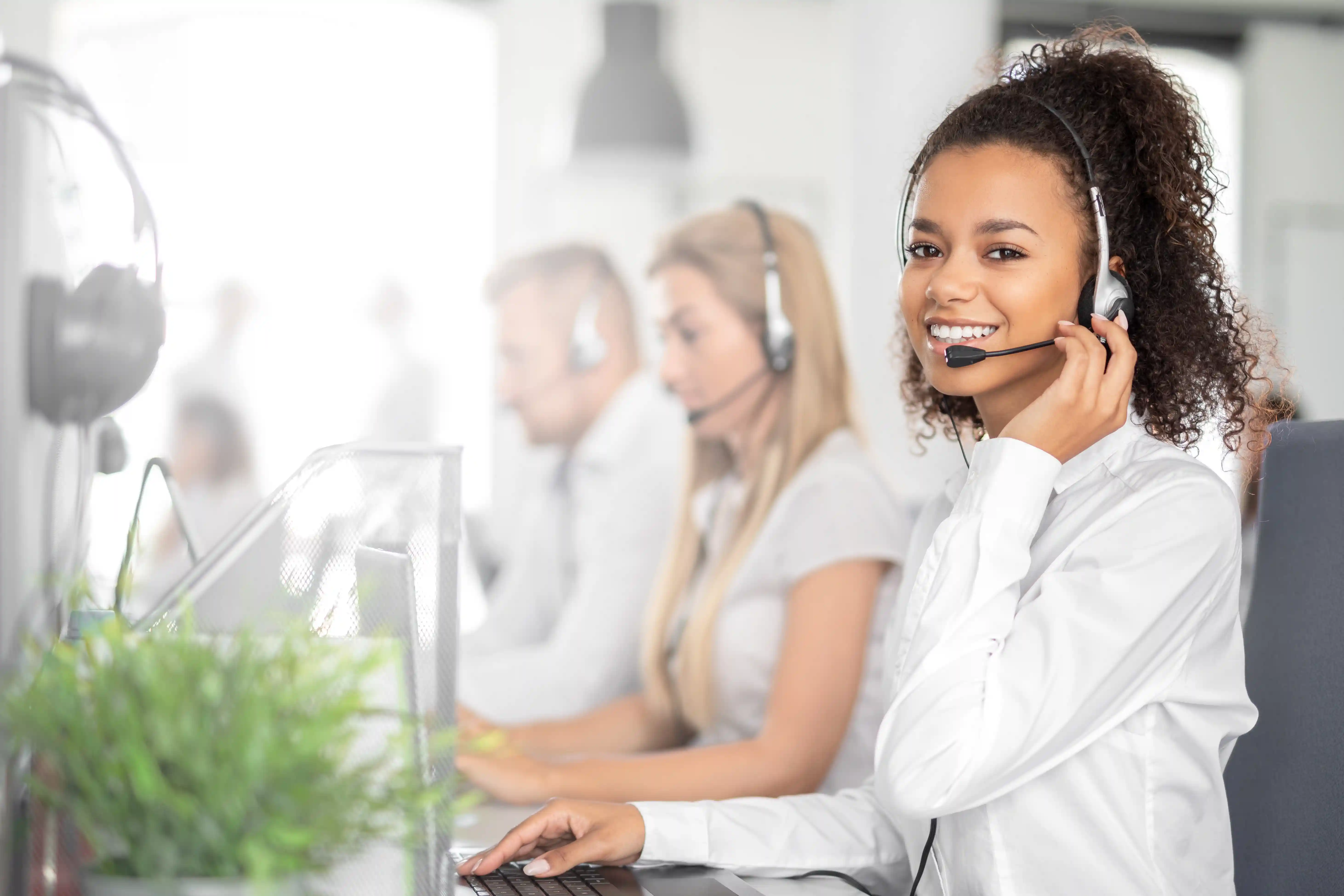 Smilende kvinde med headset arbejder i et callcenter sammen med kolleger i baggrunden – symboliserer venlig og professionel kundeservice.