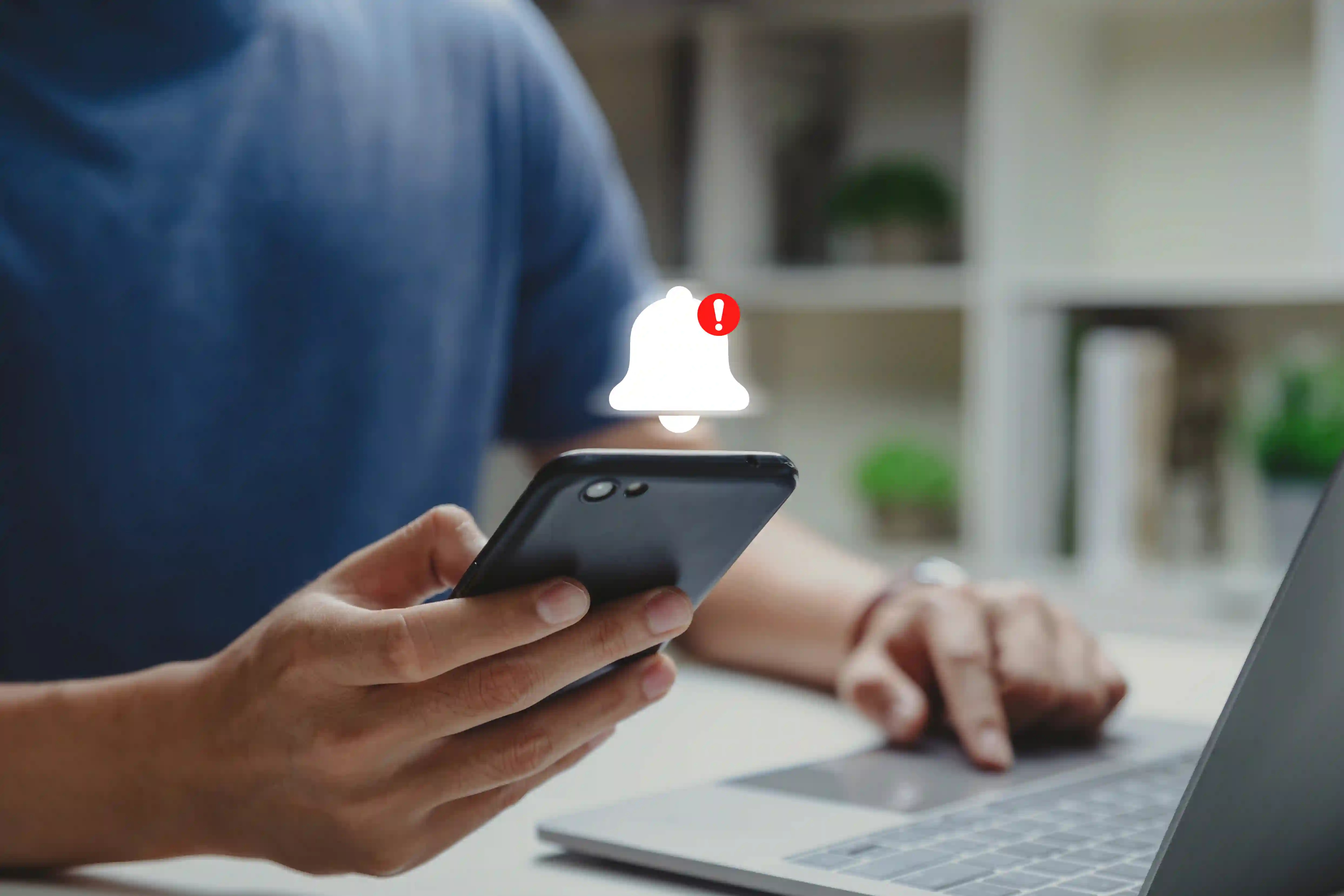 Person sidder ved en bærbar computer og holder en smartphone, hvor en notifikations-ikon med rød advarsel vises. Billedet illustrerer vigtigheden af at reagere på opkald og beskeder i tide – og hvordan Telefonpasning kan sikre, at vigtige henvendelser aldrig bliver overset.