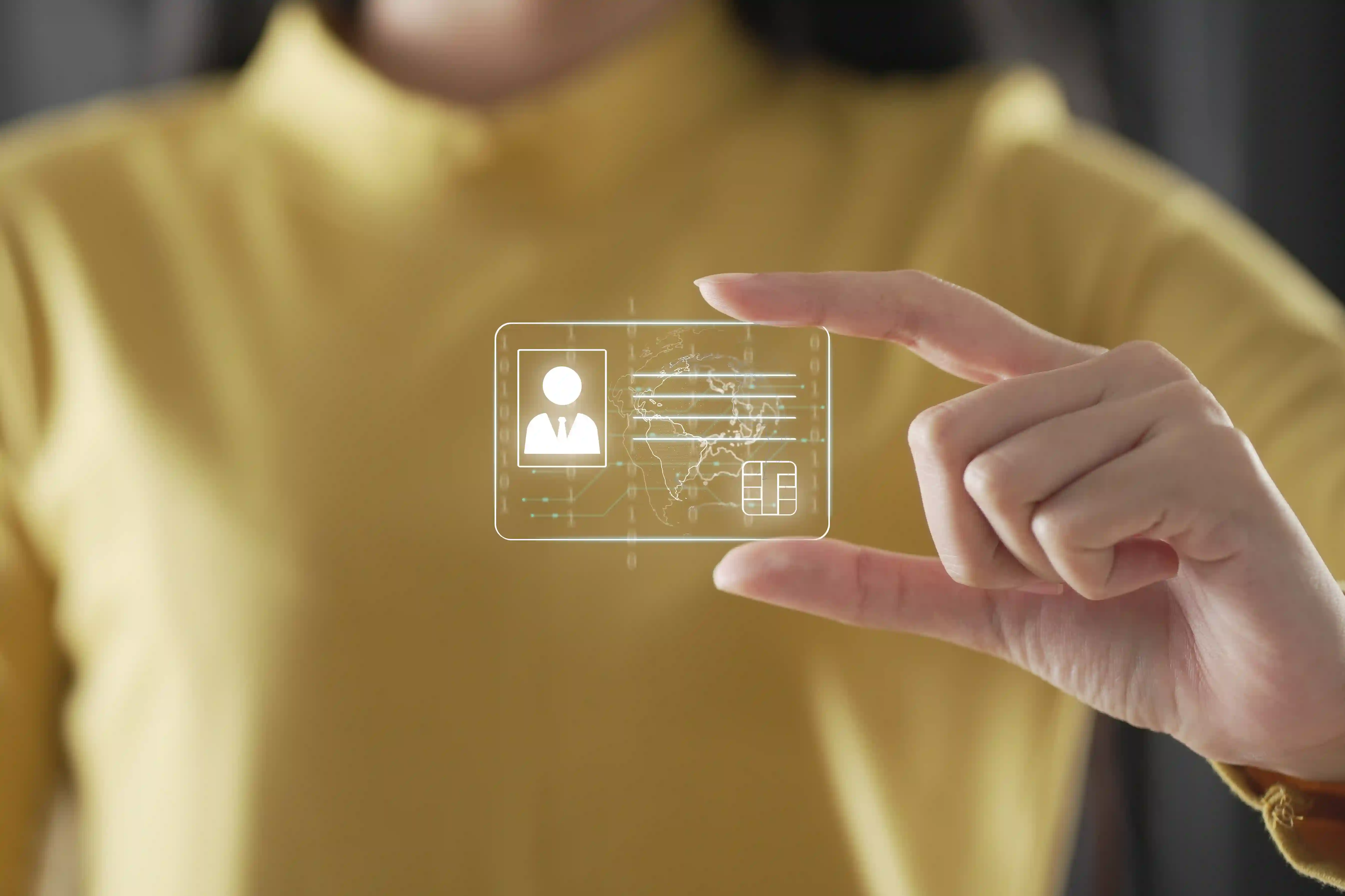 Person iført gul skjorte holder en gennemsigtig digital ID-kort-visualisering mellem fingrene. Kortet viser ikon for brugerprofil, chip og verdenskort – et symbol på digital identitet og sikker verificering, som er relevant i forbindelse med Virtuelt Kontor og KYC-processer.