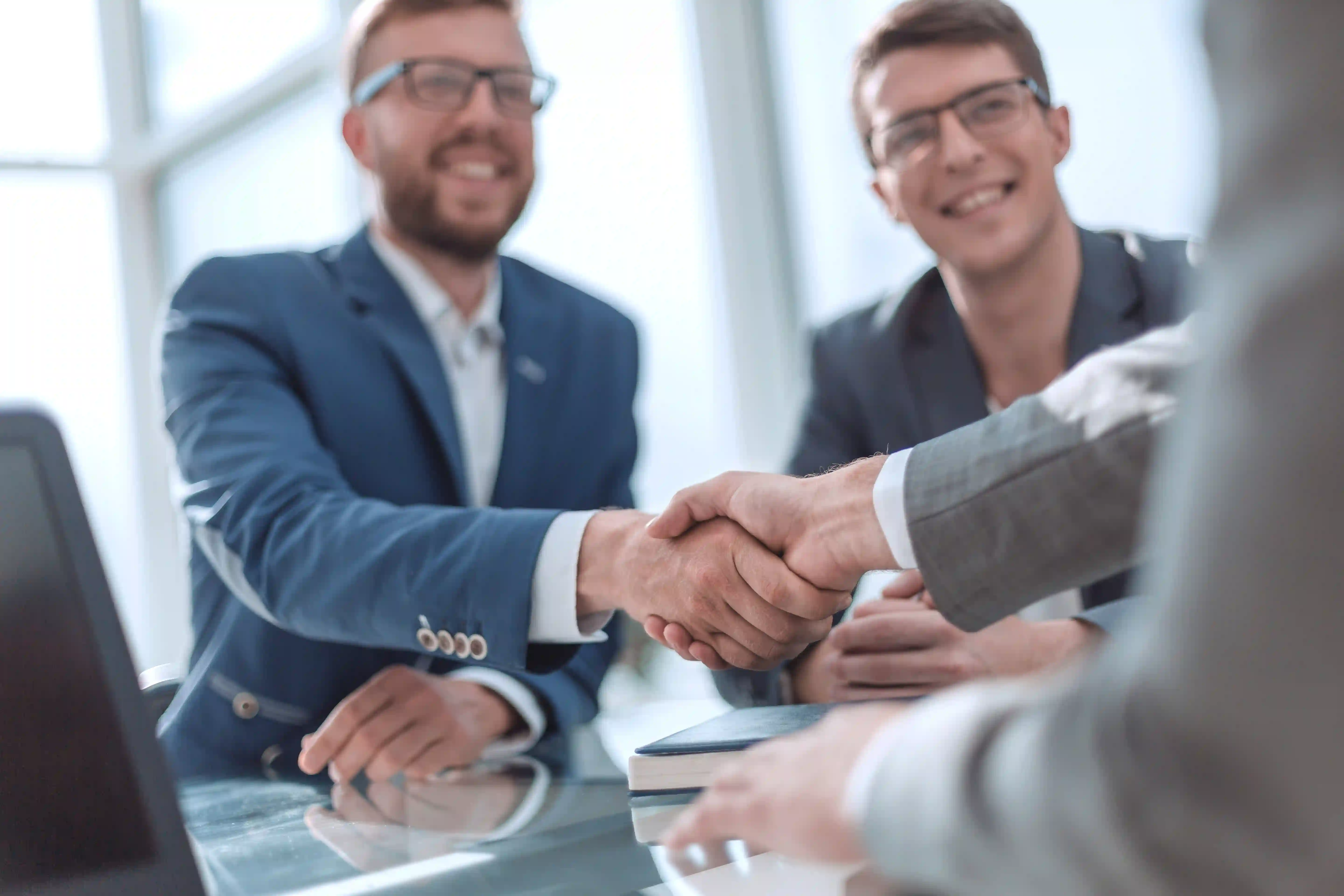 Three businessmen in suits sit at a table in a bright office environment. Two of them give each other a firm handshake, while the third smilingly monitors the encounter. The image exudes a professional atmosphere and symbolizes business agreements, partnership, cooperation or the conclusion of a successful negotiation.